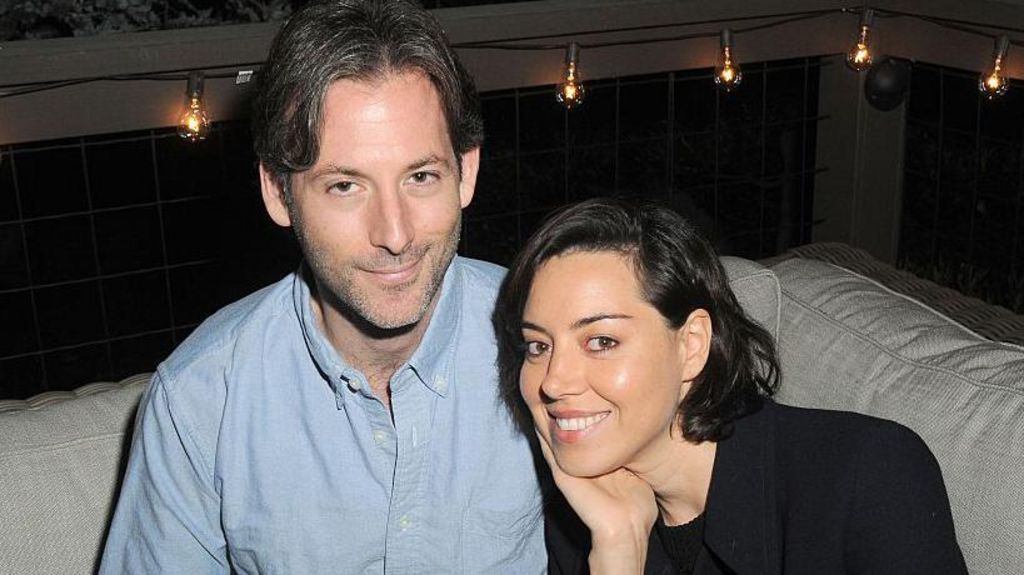 Jeff Baena and Aubrey Plaza at a party together in 2016, sitting on a sofa smiling. He is wearing a light blue shirt and has short brown hair. She has her black hair in a bob style and is wearing a black top. 