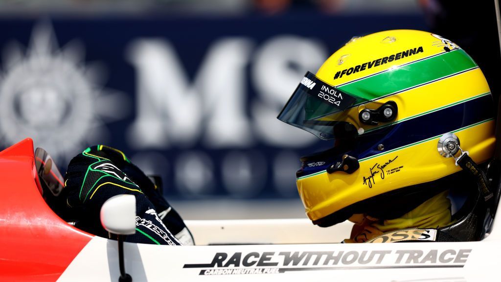 A close-up side image of Sebastian Vettel driving Ayrton Senna's 1993 McLaren while wearing a helmet with the Senna yellow, green and blue design