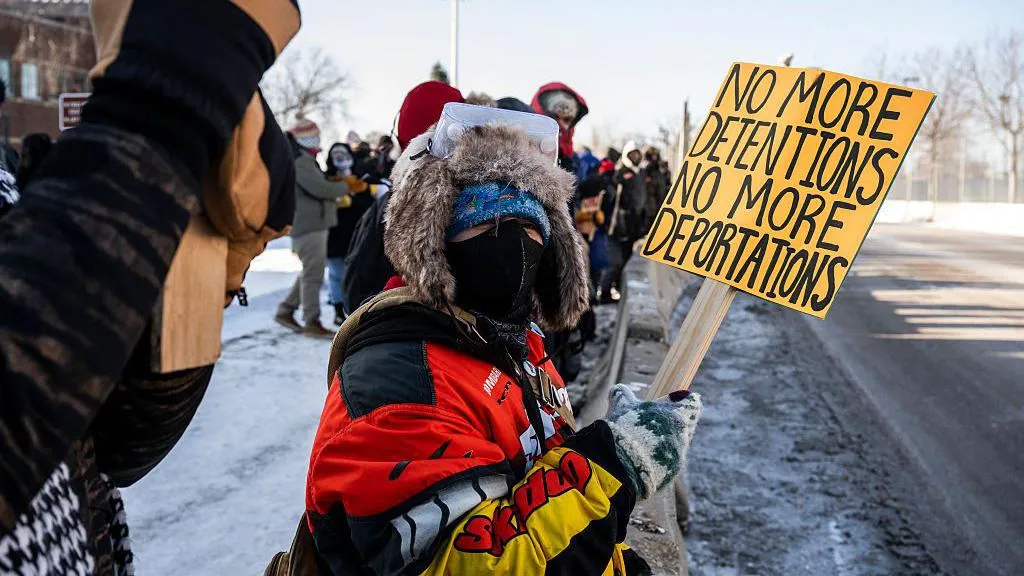Protesters outside the Bishop Henry Whipple Federal Building, near the Minneapolis-St Paul International Airport. A woman in the foreground wearing a coat and mask holds a yellow sign that reads: 