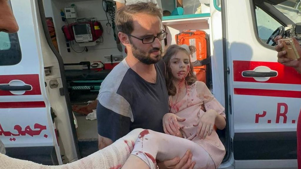 A dusty and bloodied young girl is carried from an ambulance with a heavily bandaged leg