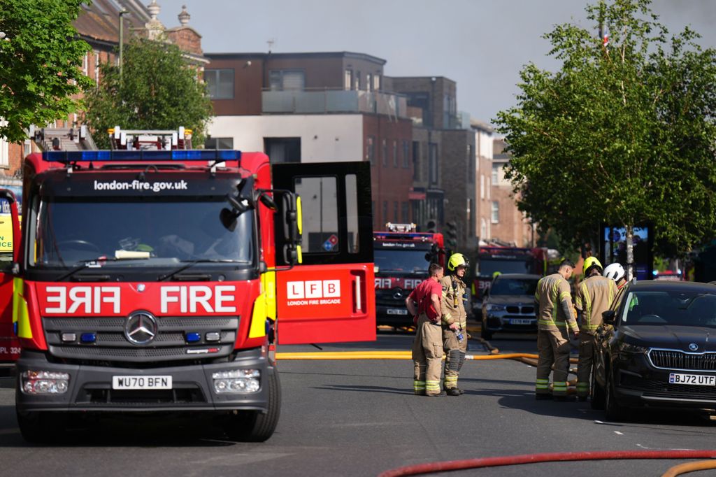 Firefighters at the scene with engines and hoses across street by parked cars