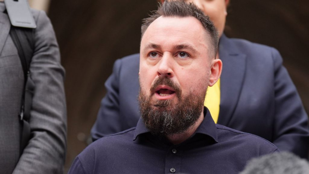 Manchester Arena attack survivor Martin Hibbert looks above the camera while talking to the media outside the High Court in London. Martin has stubble and hair which has been gelled up ever so slightly. he is wearing a navy polo shirt.