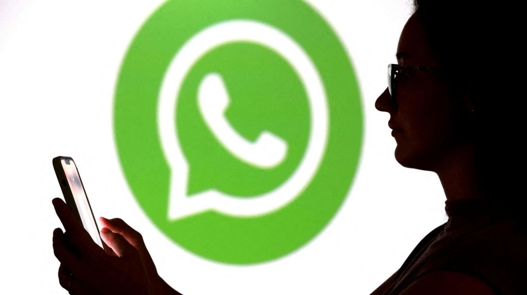 A teenager poses for a photo while holding a smartphone in front of a Whatsapp logo