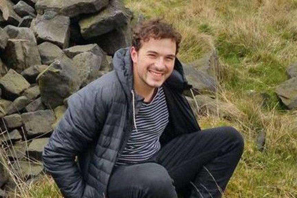 A man sits on the ground; behind him is a collapsed drystone wall and wild grass. He has short dark brown hair. He is wearing a dark blue padded coat with panels and a hood, a hooped dark blue and white top and dark jeans. He is looking at the camera, smiling