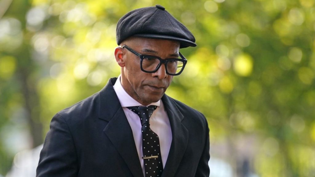 Jay Blades, wearing a dark cap, black suit and glasses looks downwards as he arrives at court 