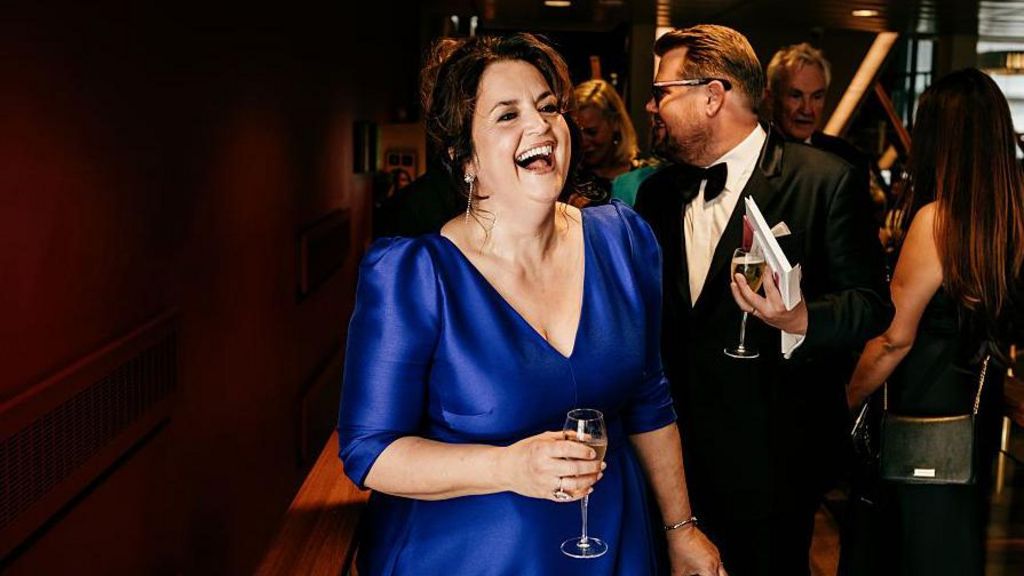 Ruth Jones in a blue dress has a giggle as she holds a glass of champagne. Her co-star and co-writer James Corden is just behind her.