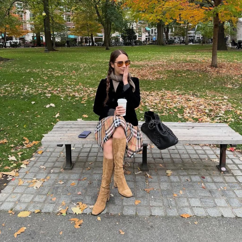 Laufey sits on a park bench in Boston. She's holding a cup of coffee, with her iphone placed beside her. Her hand is held to her face, as if in contemplation.