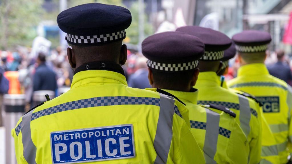 Five officers stand in a row in their yellow and blue uniforms. Each wears a black hat and stand looking out to a crowd of people. 