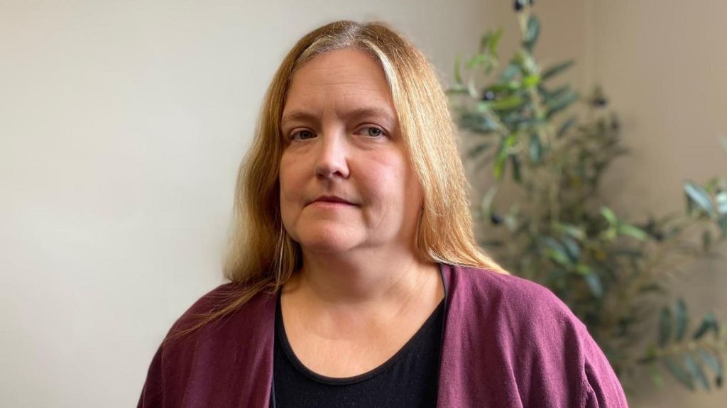 Anna has long blonde hair. She is wearing a black top and a purple cardigan. Behind her are the green leaves of an indoor plant in front of a white wall.