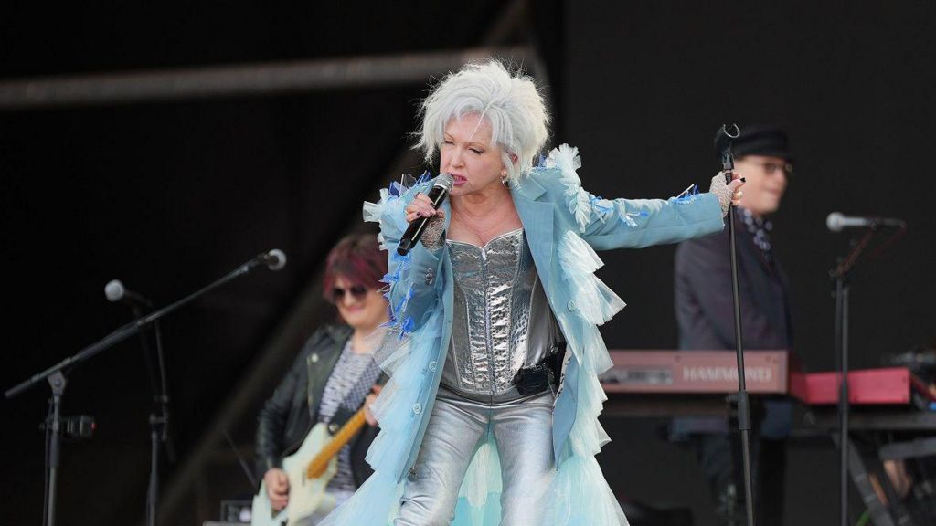 Cyndi Lauper wearing silver outfit with blue coat over while singing on stage.