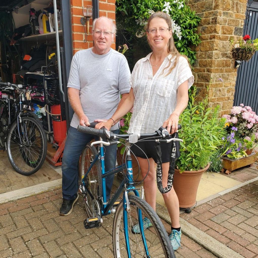 Becky wearing a white top and black shorts. She is standing with a man wearing a grey t-shirt who is holding a blue bike