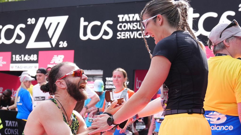 A male runner rests on one knee at the finish line, holding an open ring box as he proposes to a woman who is smiling and holding his hands.