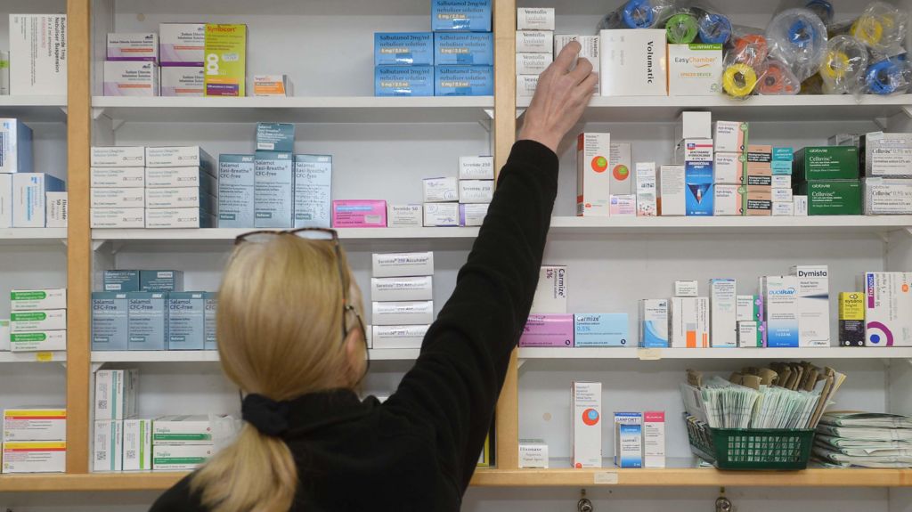The photo shows a woman with blonde hair tied in a low ponytail, with glasses on her head, wearing a black jumper. She is facing away from the camera and reaching onto the top shelf of a wall full of different medications.