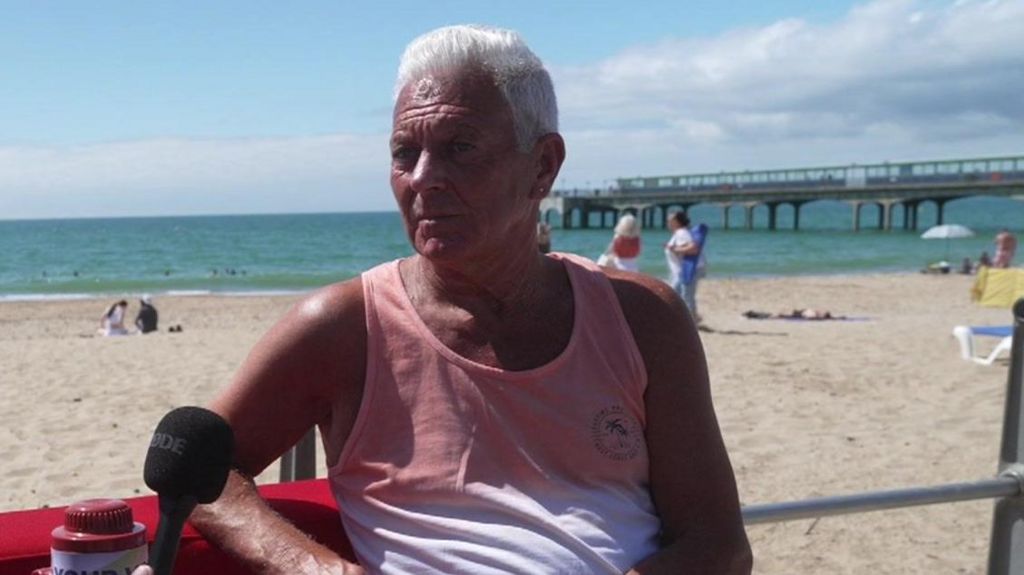 Steve is wearing a white and pink vest and has short white hair - he is sitting on a red sofa in front of Bournemouth beach