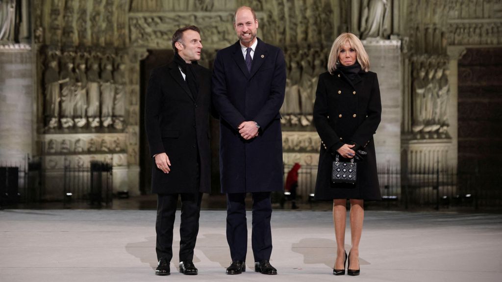 President Macron, wearing a black overcoat and scarf with a white shirt, says something to Prince William, who is also wearing a black overcoat and a white shirt with a blue tie. Brigitte Macron, wearing a black coat, black scarf and beaded black bag looks on beside them as they stand outside the Notre Dame