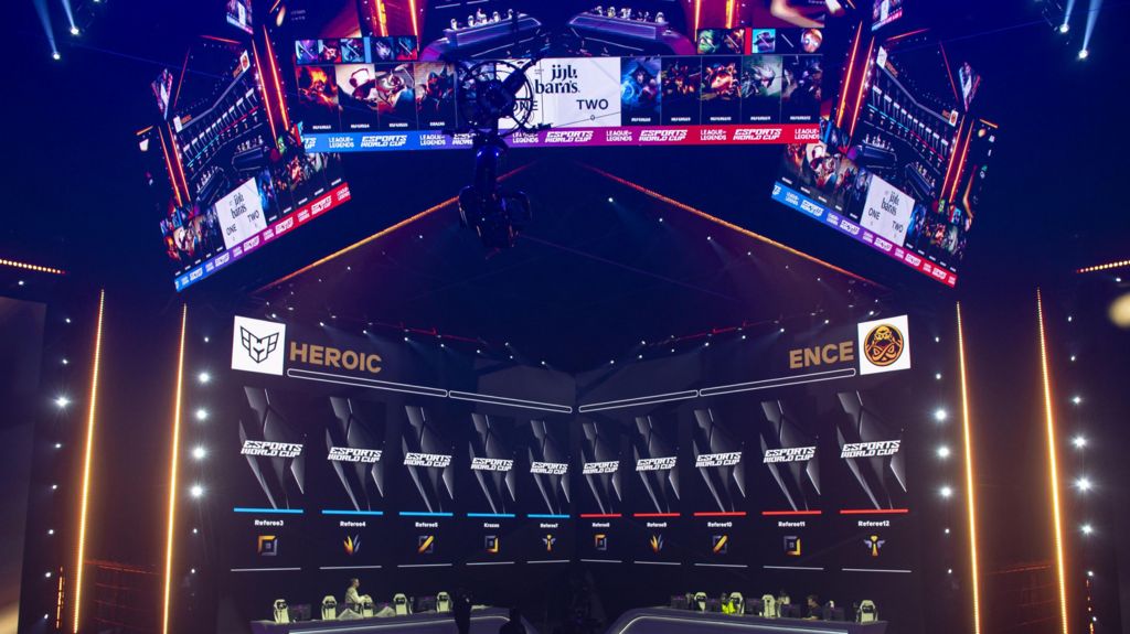 Stands are prepared for visitors during the opening ceremony of 2024 Esports World Cup in Riyadh, Saudi Arabia. The stage is lit purple with two long desks side by side in front of a score board. 