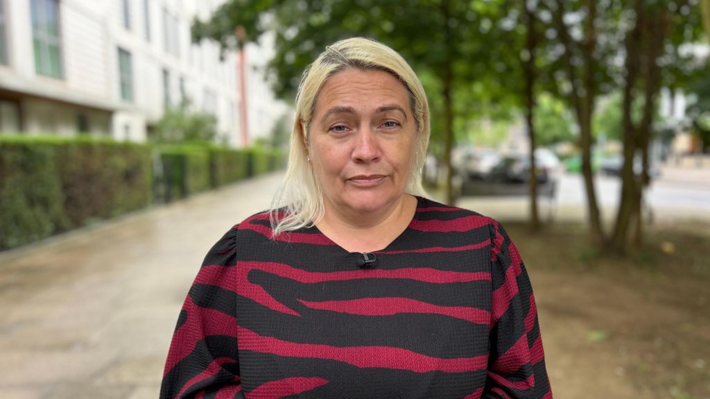A woman with light blonde hair standing outdoors on a residential street, wearing a black and red striped top, with blurred buildings, trees, and parked cars in the background