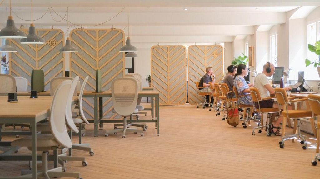 An office co-working space with floorboards, desks and wheely chairs. People in the background are working on computers with headphones on.