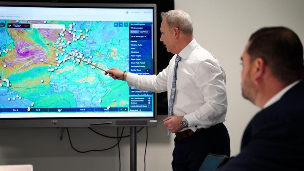 He uses a metal stick to point at a weather map on a screen. Another man in a black suit sits in the foreground watching. 