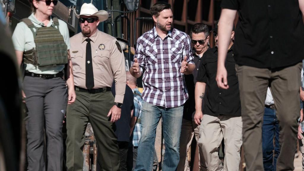 JD Vance in red-checked shirt and jeans walks with border officials