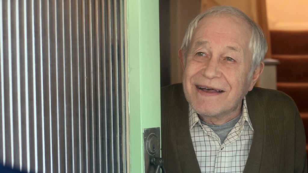 An older Rothwell wearing a shirt and cardigan and opening a door in character during a scene from Doctors.