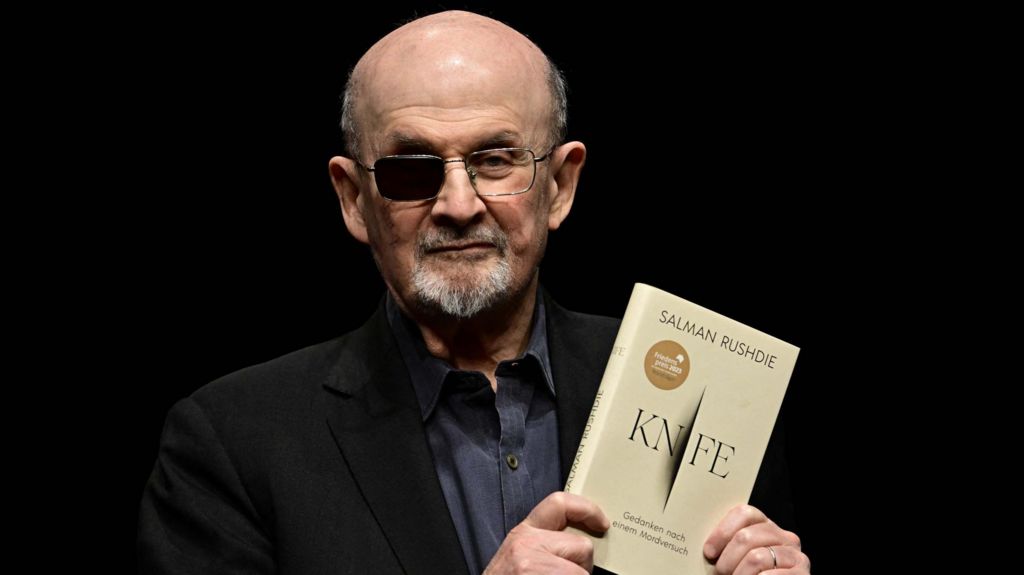 Salman Rushdie wearing a black jacket and black shirt, holding up his book Knife. He is wearing glasses with the right lens blacked out.