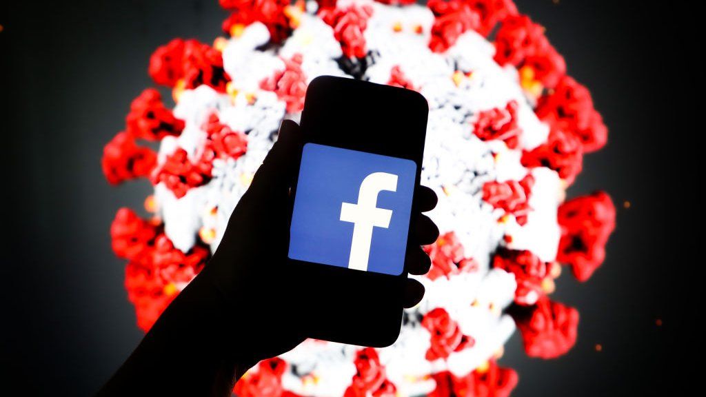 Facebook logo is displayed on a mobile phone screen photographed on SARS-CoV-2 illustration graphic background.