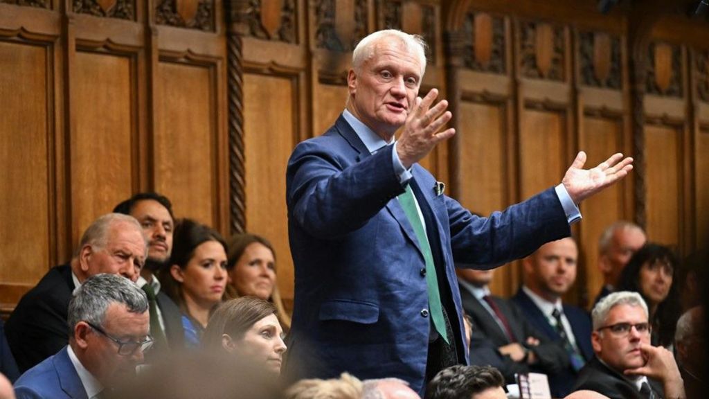 Graham Stuart standing in the Commons wearing a blue suit and green tie with his arms raised, with other MPS in the background