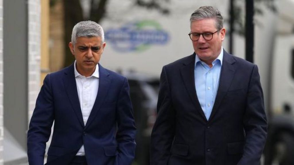 Sadiq Khan and Keir Starmer walk side by side down a pavement, both wearing dark suits. A brick building runs along one side of the pavement and a lorry is visible in the background.