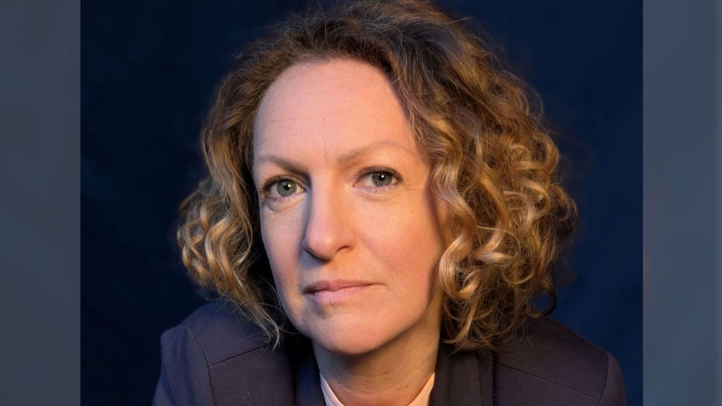 A professional headshot of Victoria Woodall with chin length golden curly hair wearing a navy suit against a navy backdrop