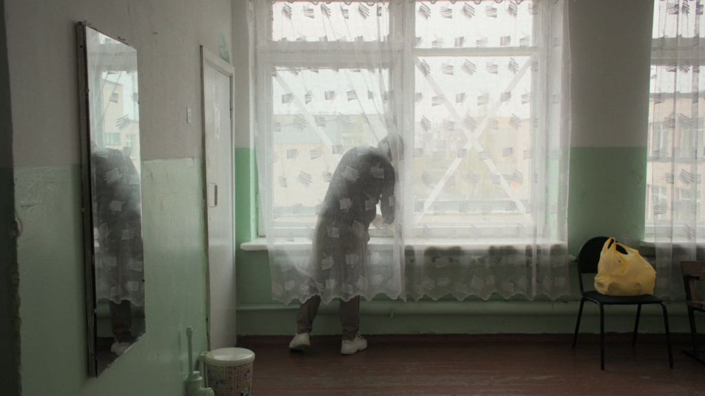 Pasha behind a net curtain sticking an X symbol on the window. chair with yellow bag. On left hand side a mirror. Green walls
