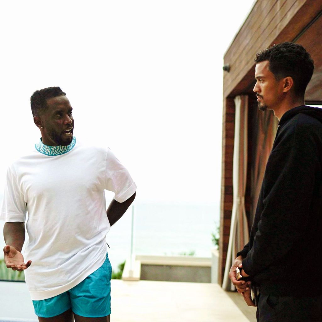 Sean Combs (left) and Phil Pines (right) talk outdoors near a building with an ocean view in the background; Combs wears a white t-shirt and blue shorts, Pines is in a black hoodie.