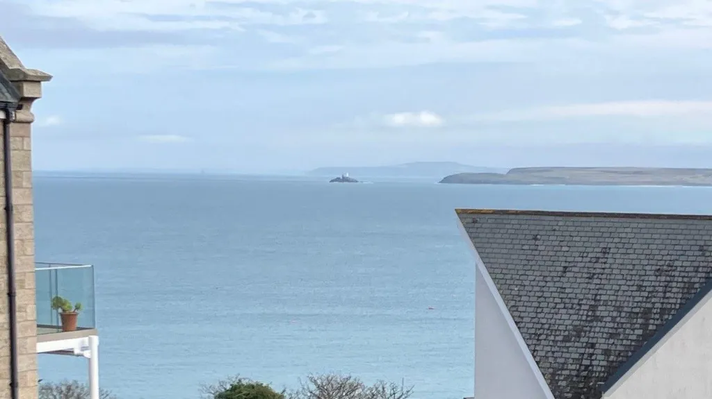 The view from Virginia Woolf's St Ives holiday home across the bay to Godrevy Lighthouse and the coast beyond it on a clear day. Either side of the view are roofs and to the left is a balcony.