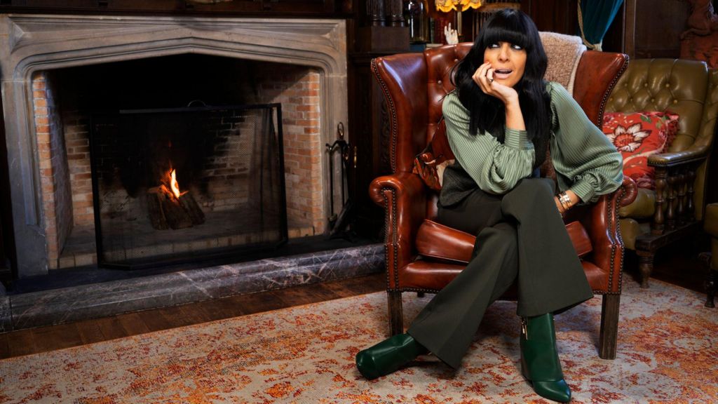 Claudia Winkleman sitting in a chair by an open fireplace