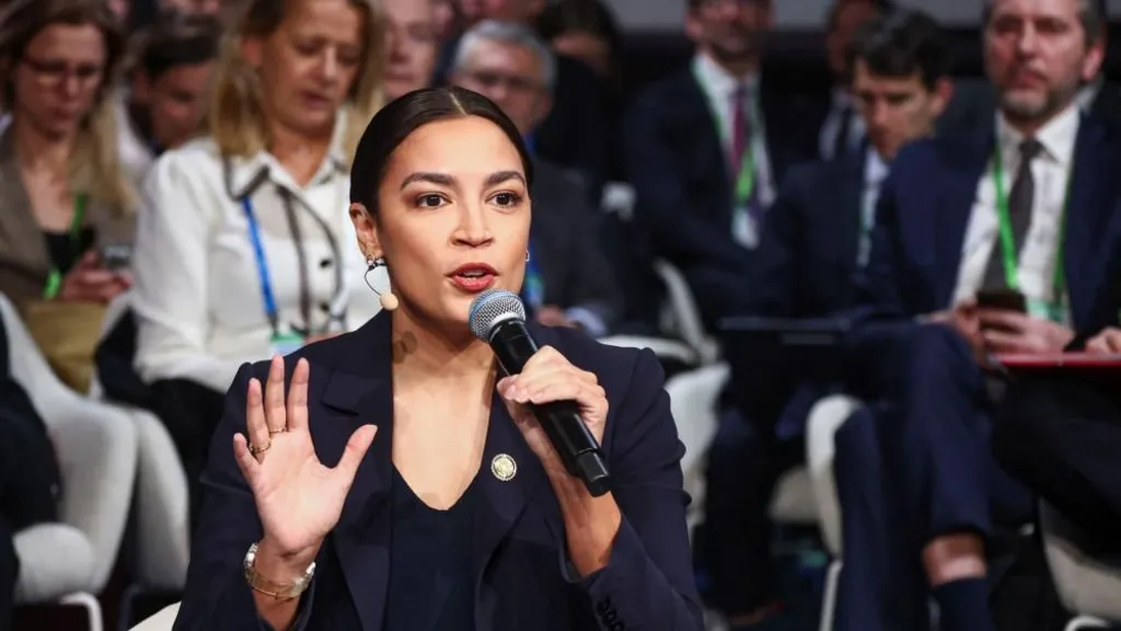 Rep Alexandria Ocasio-Cortez, wearing a dark suit and with her hair tied back, holds a microphone in one hand and gestures with an open palm with the other in front of a crowd of seated people
