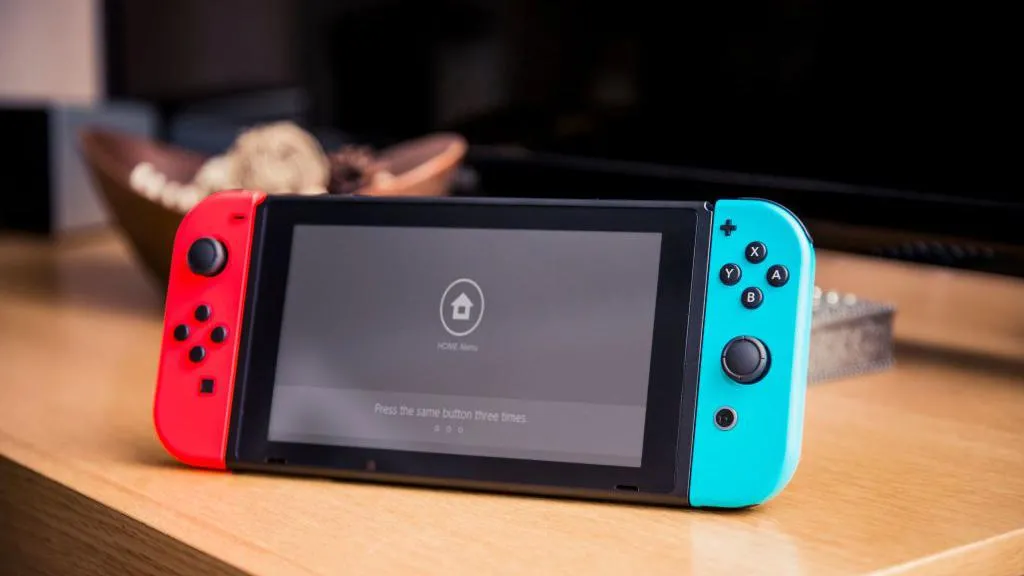 A Nintendo Switch console with red and blue joy con controllers attacked propper up on a desk. The screen is the home screen and says