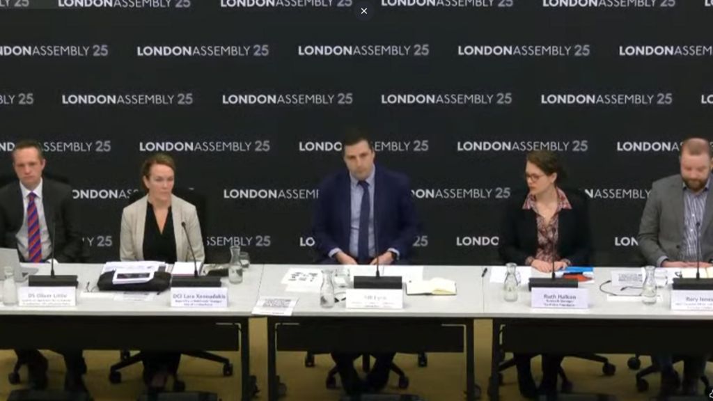 Five panel members, including three men and two women, sit behind a row of desks at a meeting. Behind them is a backdrop that says London Assembly 25.