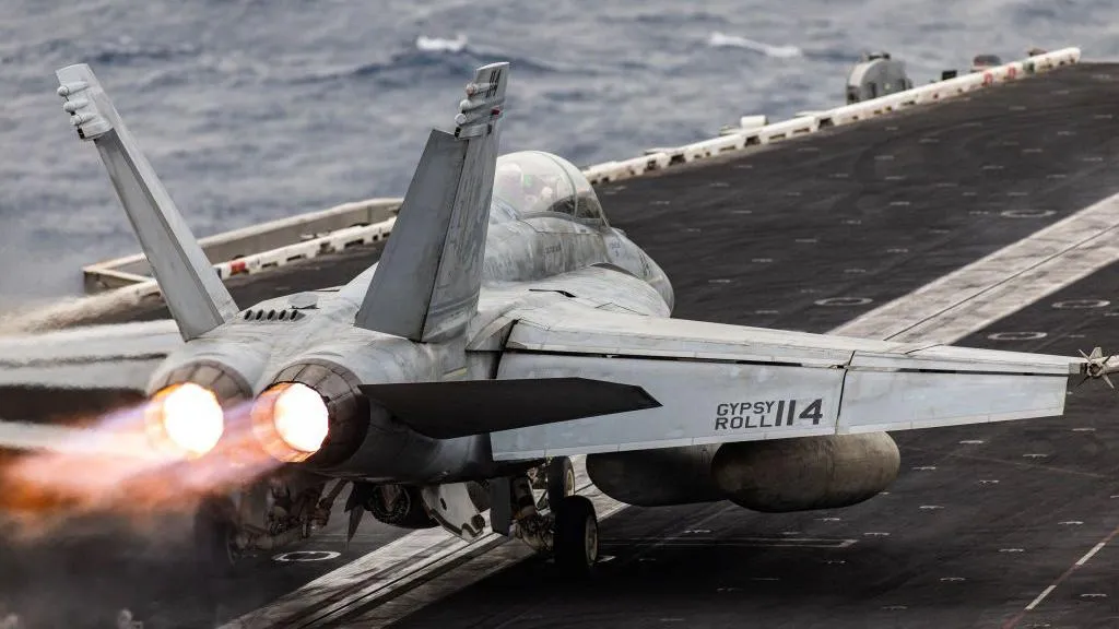 Un avión de combate despegando desde la cubierta de un portaaviones, con los motores a reacción encendidos y llamas visibles, mientras el océano se extiende en el fondo.