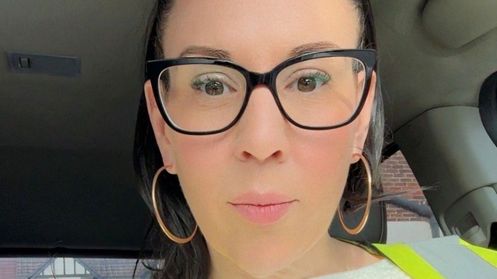 Natalie Kimber wearing glasses, big hoop earrings, sitting in a car, wearing a high-vis vest.