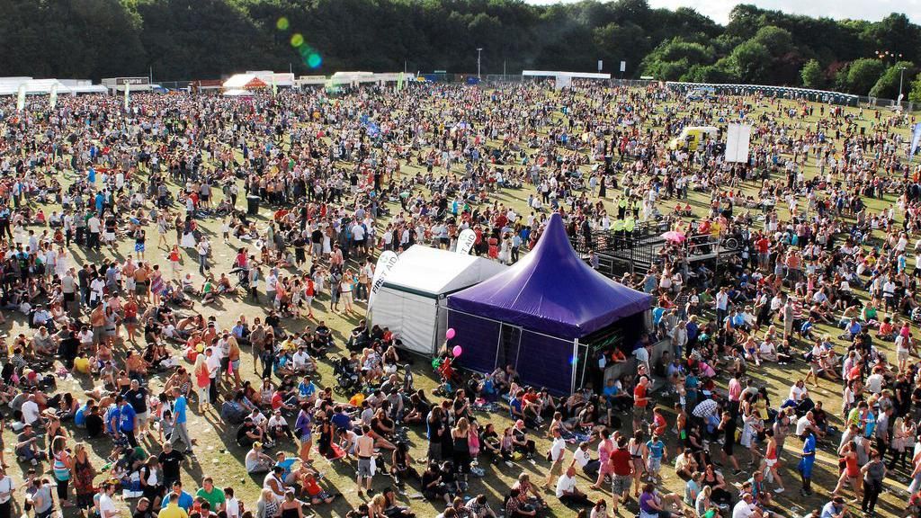 A large crowd in a field at the festival and in the centre are two tents, one a purple tent houses a stage while behind it is a white tent. Further tents are visible in the distance at the edge of the crowd