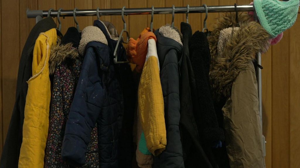 A clothes rack is full of coats with some yellow and blue ones standing out. There is a green hat hanging from the end of the rack and a brown coat with a furry hood next to it.