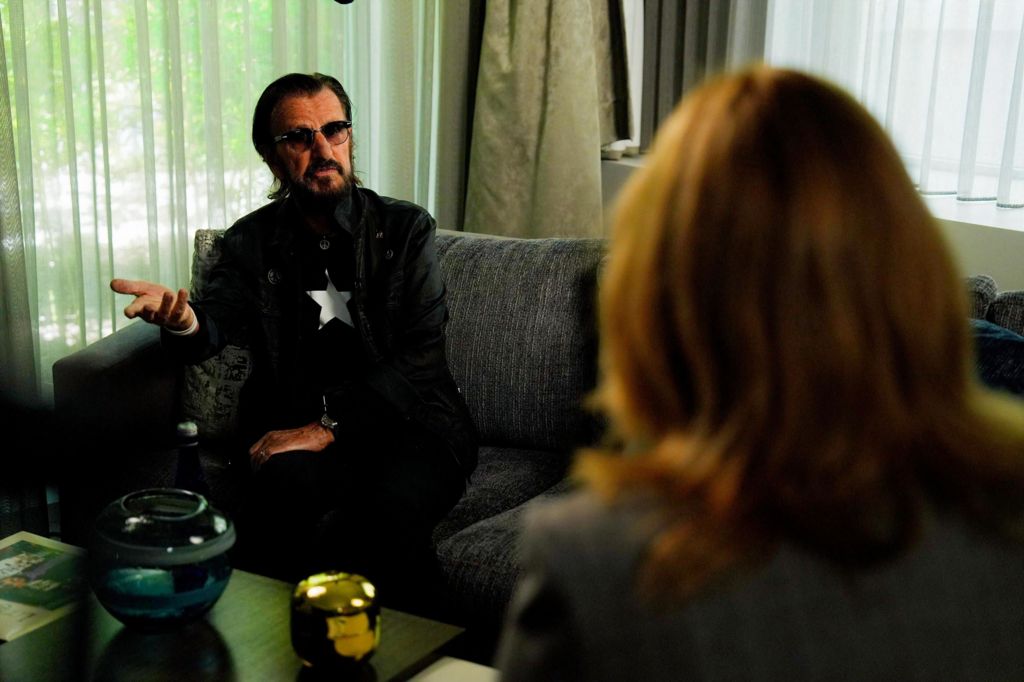Ringo Starr in the foreground as he sits on the couch speaking to a woman whose back is to the camera.