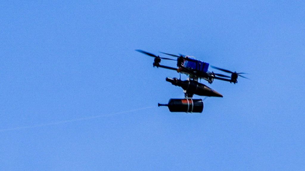 A fibre-optic drone flies in a clear blue sky across the border from Lebanon into Israel. It has a spool of fibre optic wire handing under a chassis, supported by four propeller blades around a blue rectangular battery.
