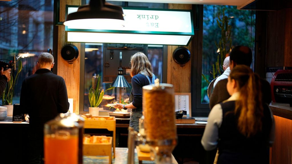 A food serving counter apparently in a hotel or catering facility and a queue of people, men and a woman, seen from behind.