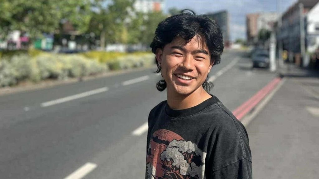 Mr Tamang, wearing a black T-shirt with a faded image on the front, is standing by the side of a road and smiling.