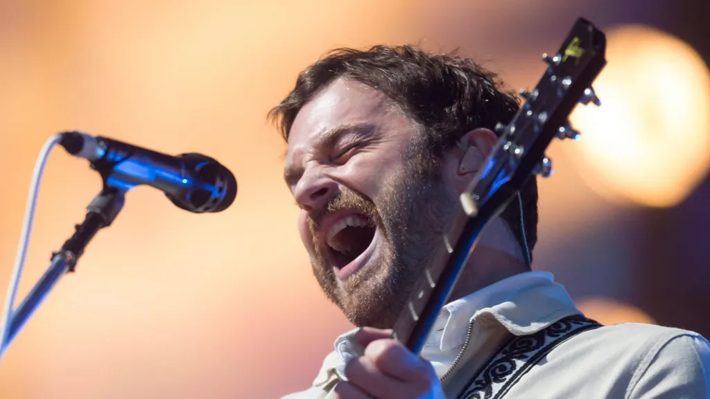 Kings of Leon lead vocalist Caleb Followill closes his eyes and sings into a microphone. He is wearing a light-coloured shirt and also playing a guitar.