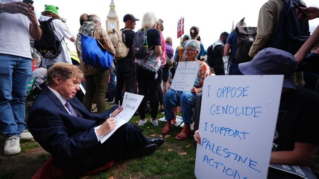 A man in a suit writes on a placard, while others sit near him holding signs