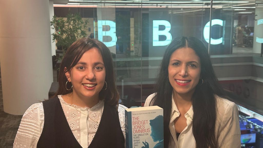 Yasmin Rufo and Noor Nanji holding a copy of the book Bridget Jones's Diary, with the BBC logo on a wall behind them