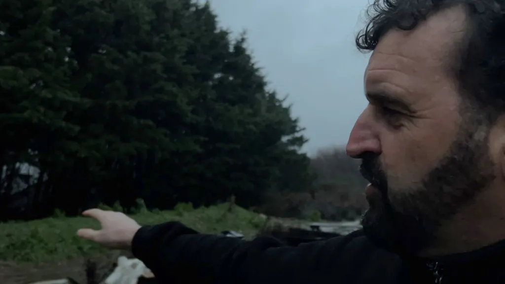 Lawrence Bates is in the right of the frame looking out to the left, where a treelined is visible. It is overcast, and Lawrence is wearing black and has a black beard and black hair. 