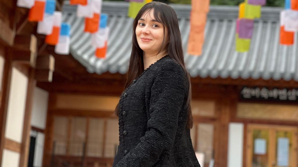 Mary Gedda, dressed in black, smiles in a photo taken in Seoul. A wooden building with a tin roof can be seen behind her